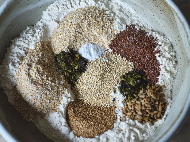 Large mixing bowl with bread ingredients including flax seeds, sunflower seeds, quinoa, flour, and bran