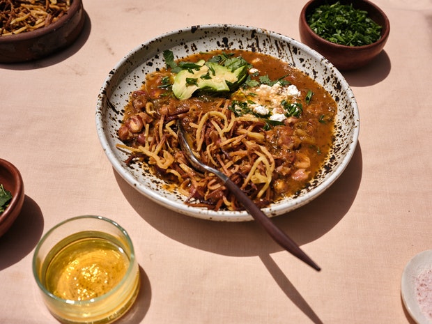 A Close-up of a bowl of Vegan Pozole with toppings including Tortilla Chips, Avocado and Cilantro